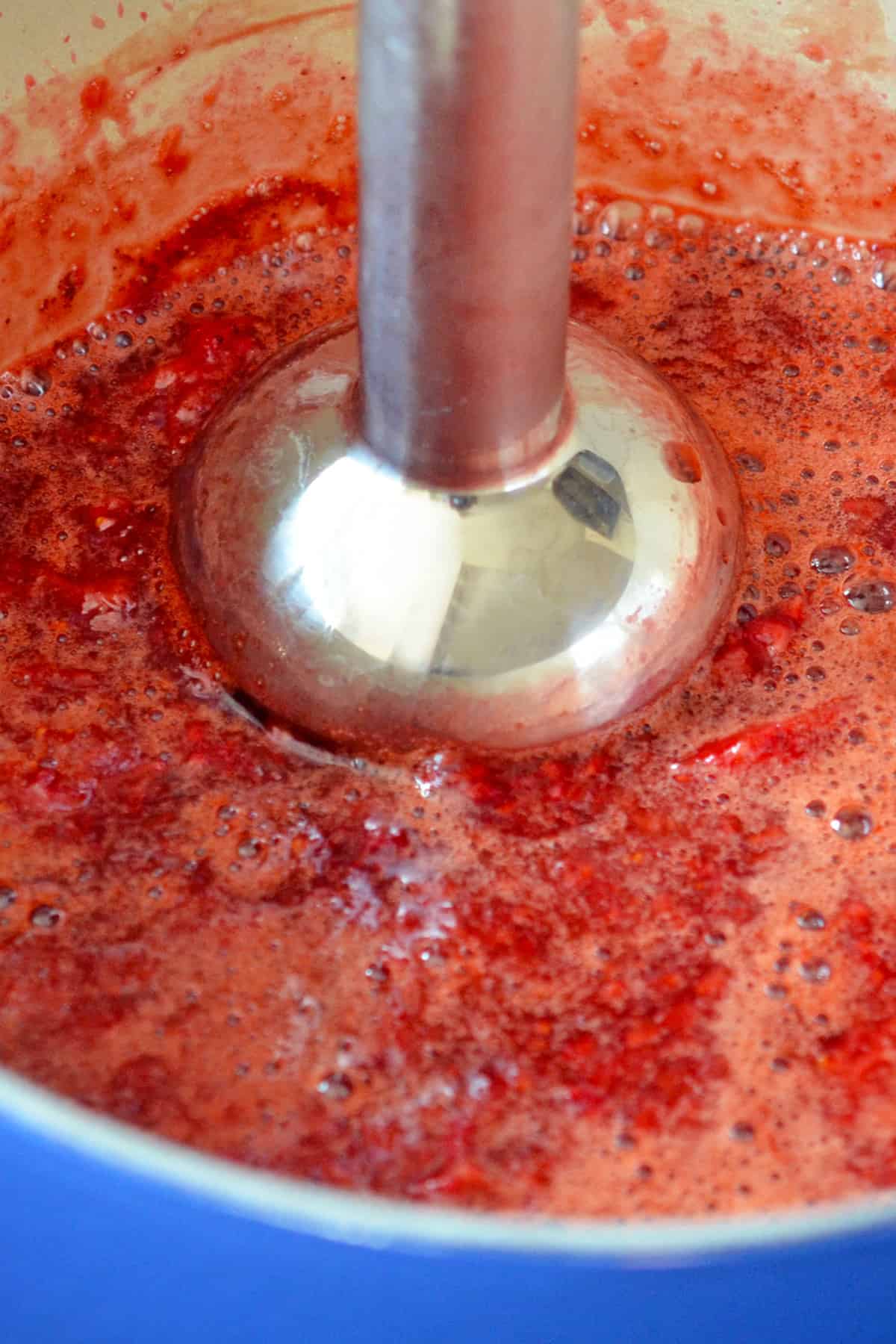 An immersion blender is pureeing a strawberry sauce in a blue saucepan.