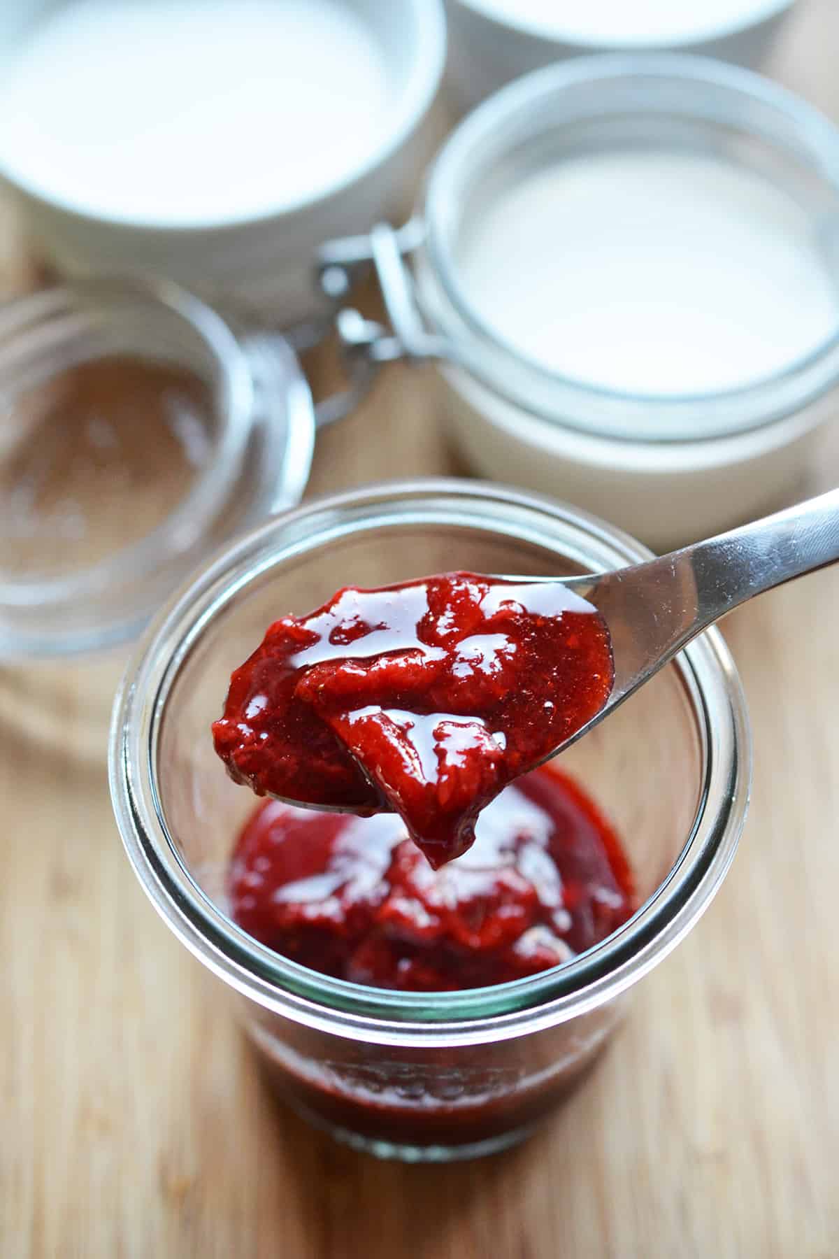 A spoonful of strawberry compote from a glass jar filled with it.
