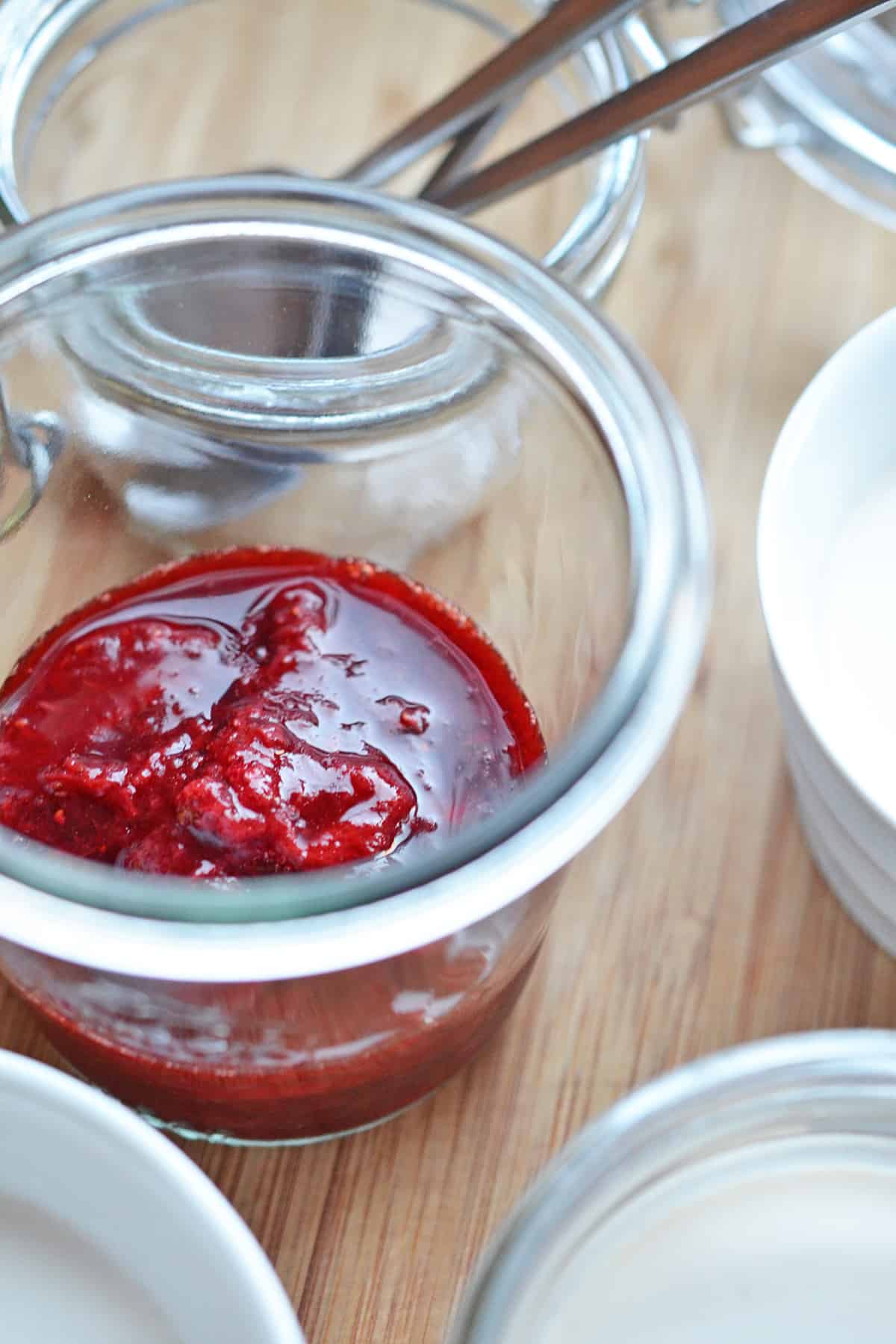 A closeup shot of a jar of homemade strawberry compote next to ramekins filled with Panna Cotta.