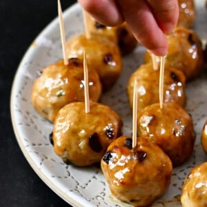 A hand is grabbing a cranberry turkey meatballs topped with a gluten free maple Dijon mustard glaze with a toothpick from a white plate.