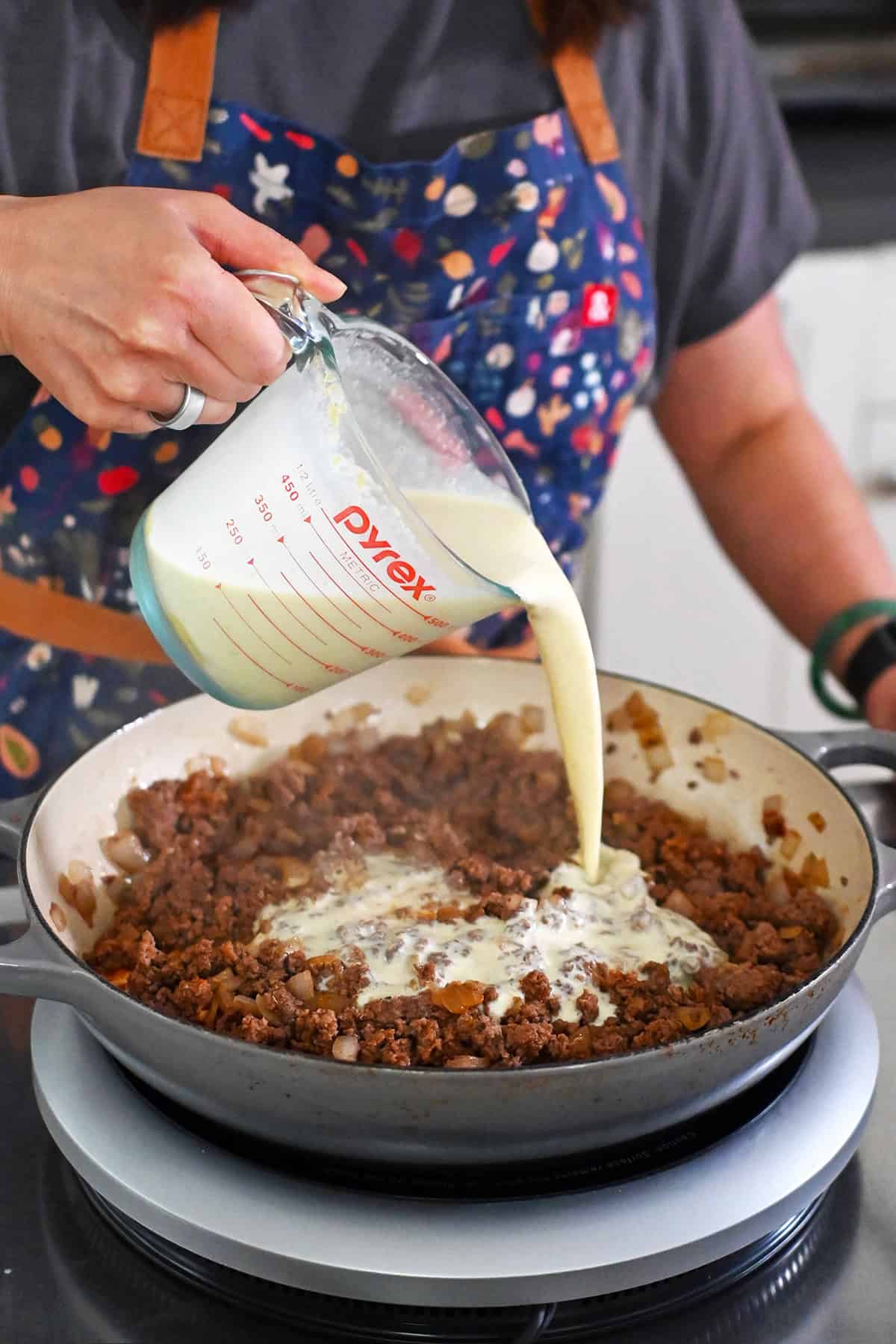 Pouring the dairy-free sauce into a large skillet filled with cooked and seasoned ground beef and diced onions.