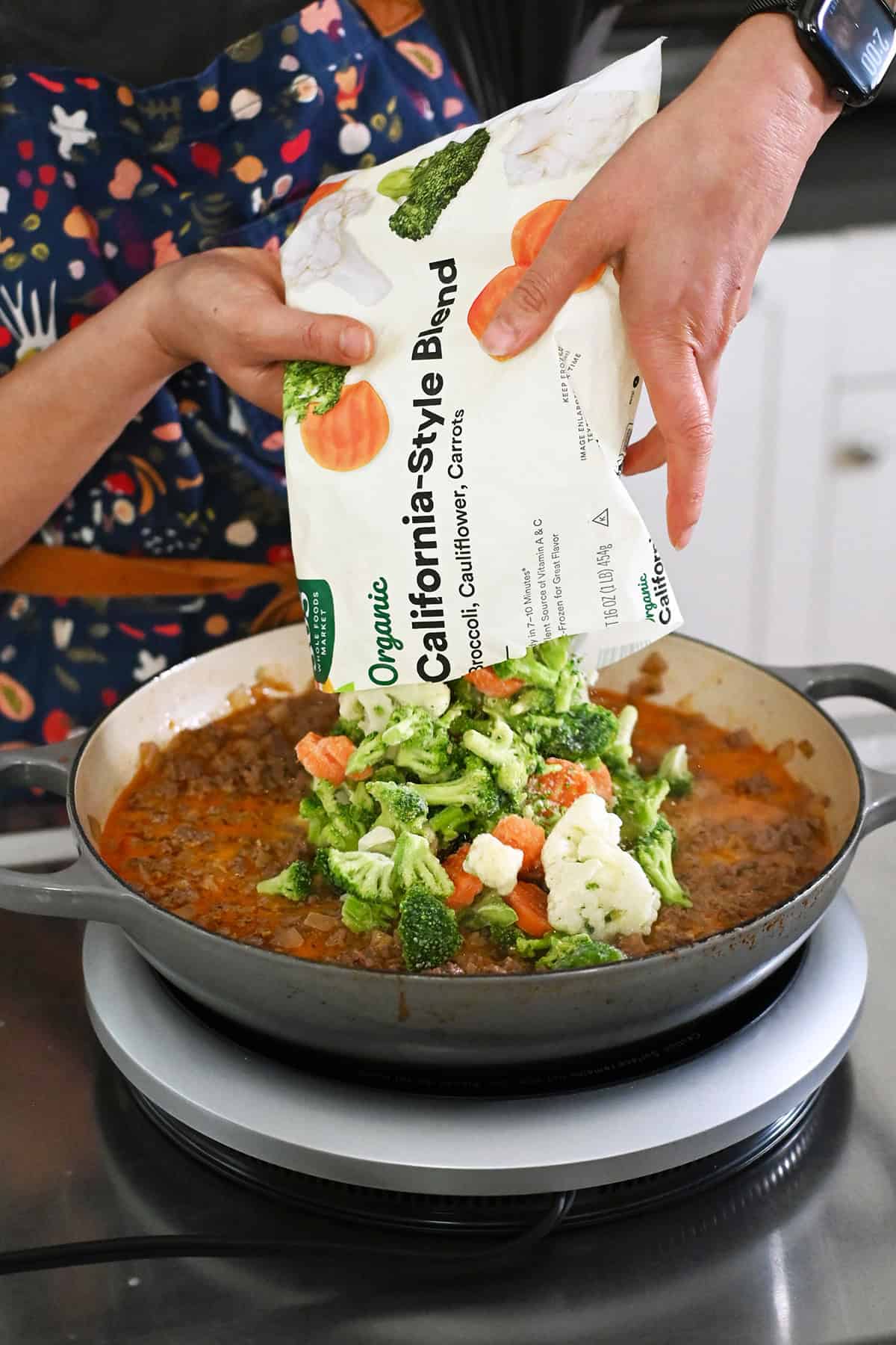 Adding an open package of frozen mixed vegetables into the skillet with ground beef, onions, and a dairy-free sauce.