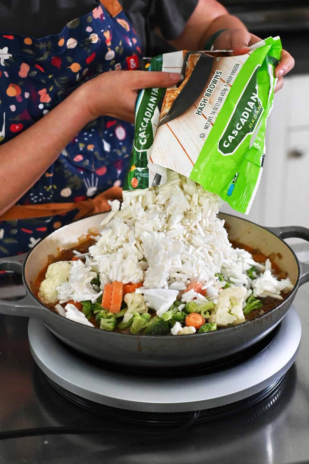 Adding an open package of frozen hash browns into the skillet with seasoned ground beef, diced onions, dairy-free sauce, and mixed frozen vegetables.