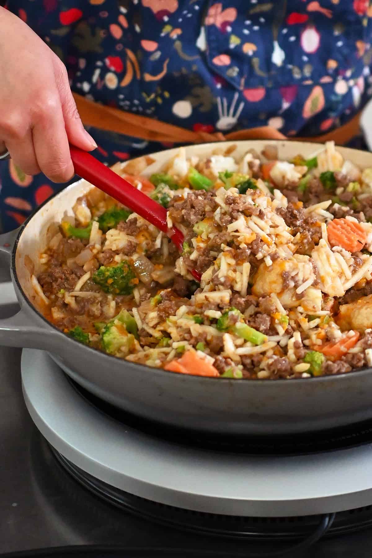 Mixing the contents of the skillet well with a red silicone spatula to integrate the frozen vegetables with the sauce and beef.