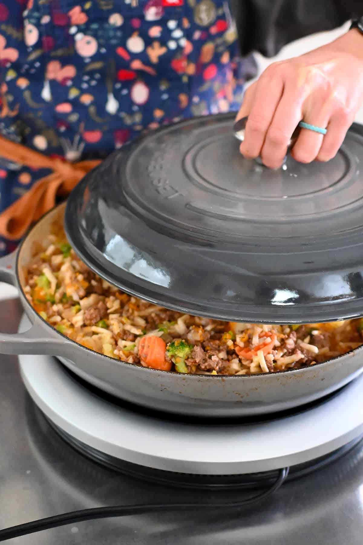 Placing a lid on top of a skillet filled with ground beef, potatoes, and frozen vegetables so it can cook evenly.
