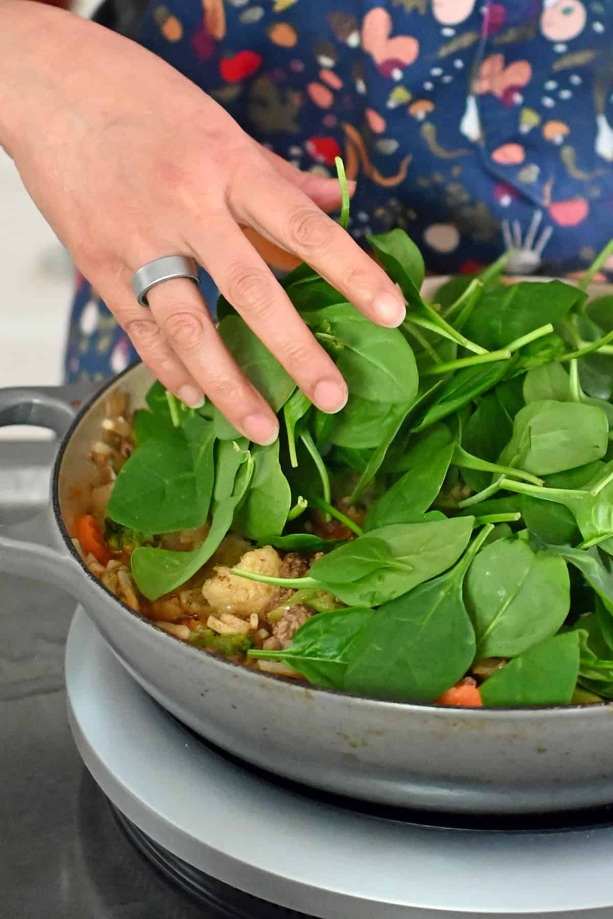Adding fresh baby spinach to a skillet filled with cooked ground beef, potatoes, and mixed vegetables.