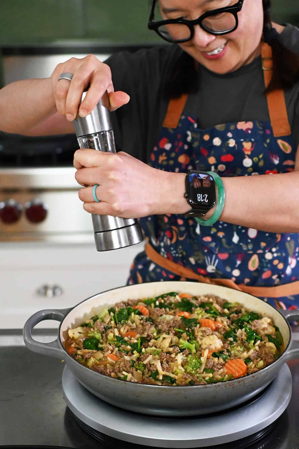 A smiling Asian woman in glasses and braids is adding freshly cracked black pepper from a pepper mill into a large gray skillet with cooked ground beef, shredded potatoes, and mixed vegetables.