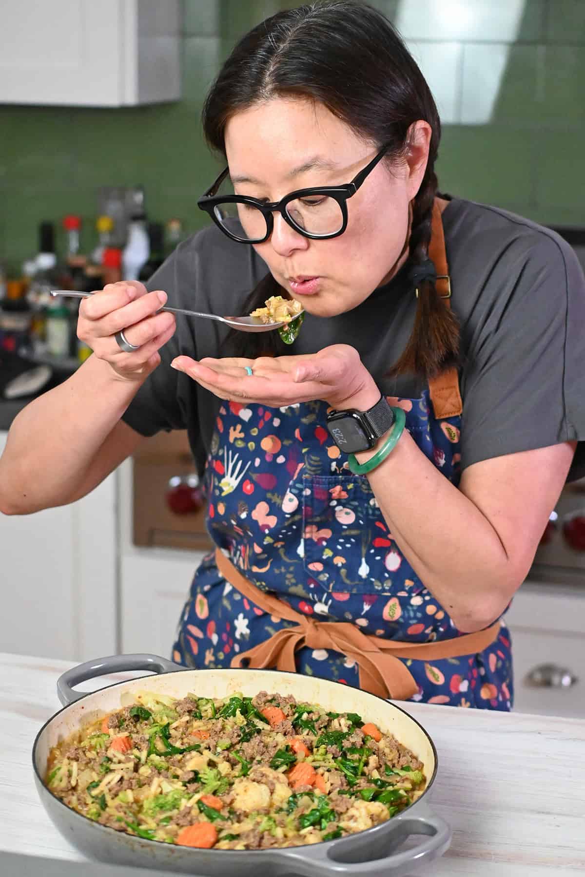 An Asian woman in glasses and braids is tasting a bite from a skillet filled with ground beef, shredded potatoes, and veggies.