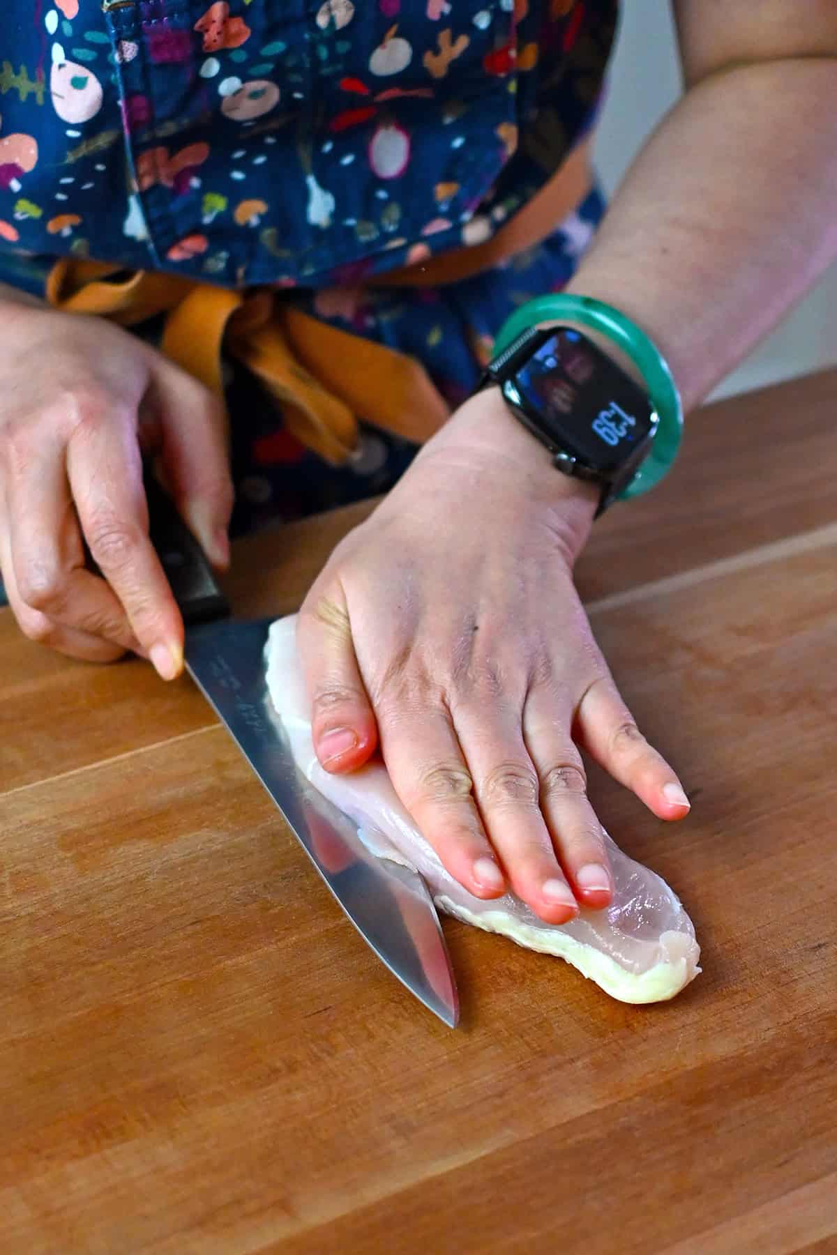A person's hand is resting flat on top of a raw boneless skinless chicken breast as they use their other hand to hold a chef's knife to slice the breast in half horizontally.