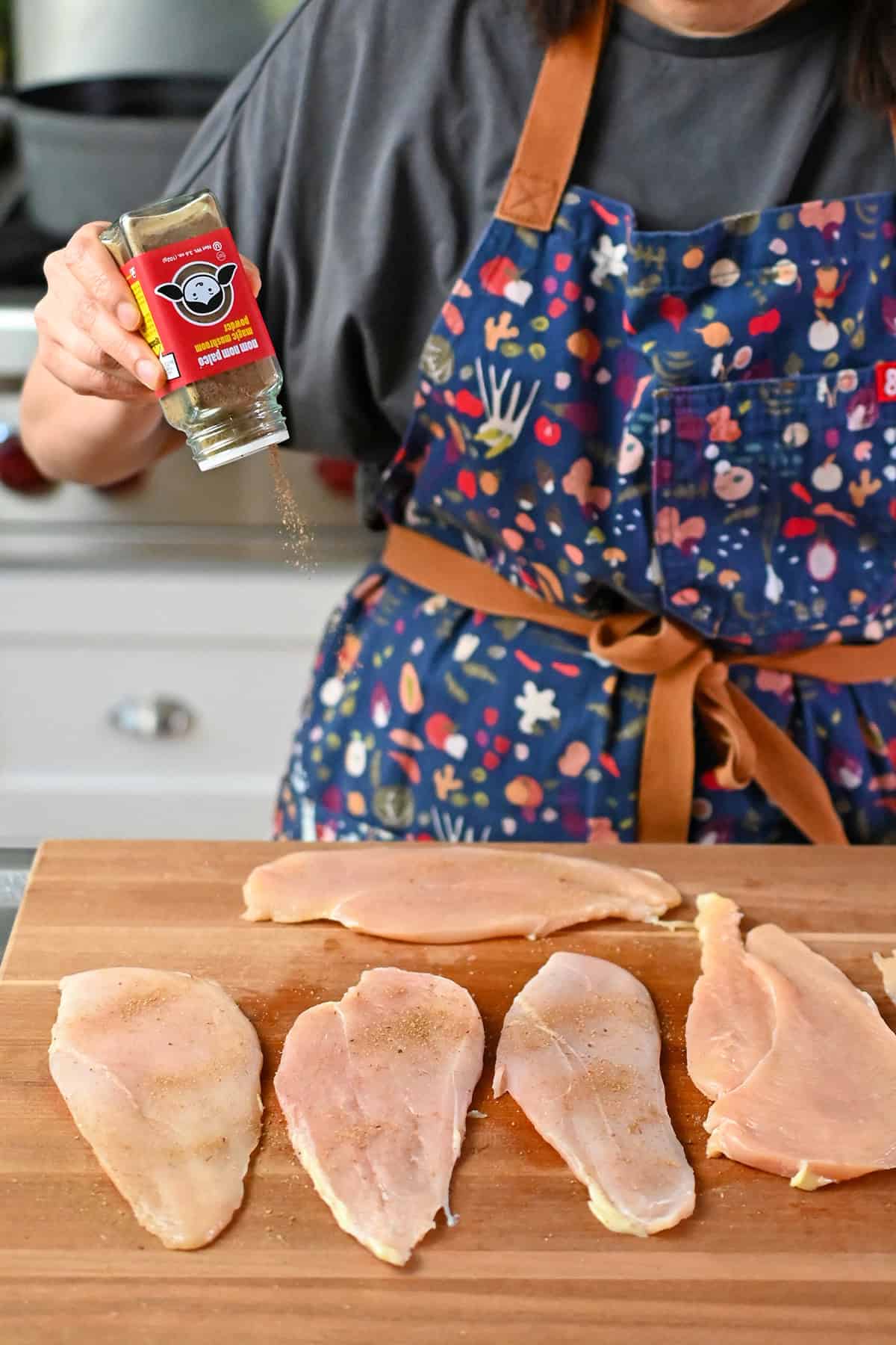 Sprinkling Nom Nom Paleo's Magic Mushroom Powder onto thinly pounded raw chicken breast cutlets on a wooden cutting board.