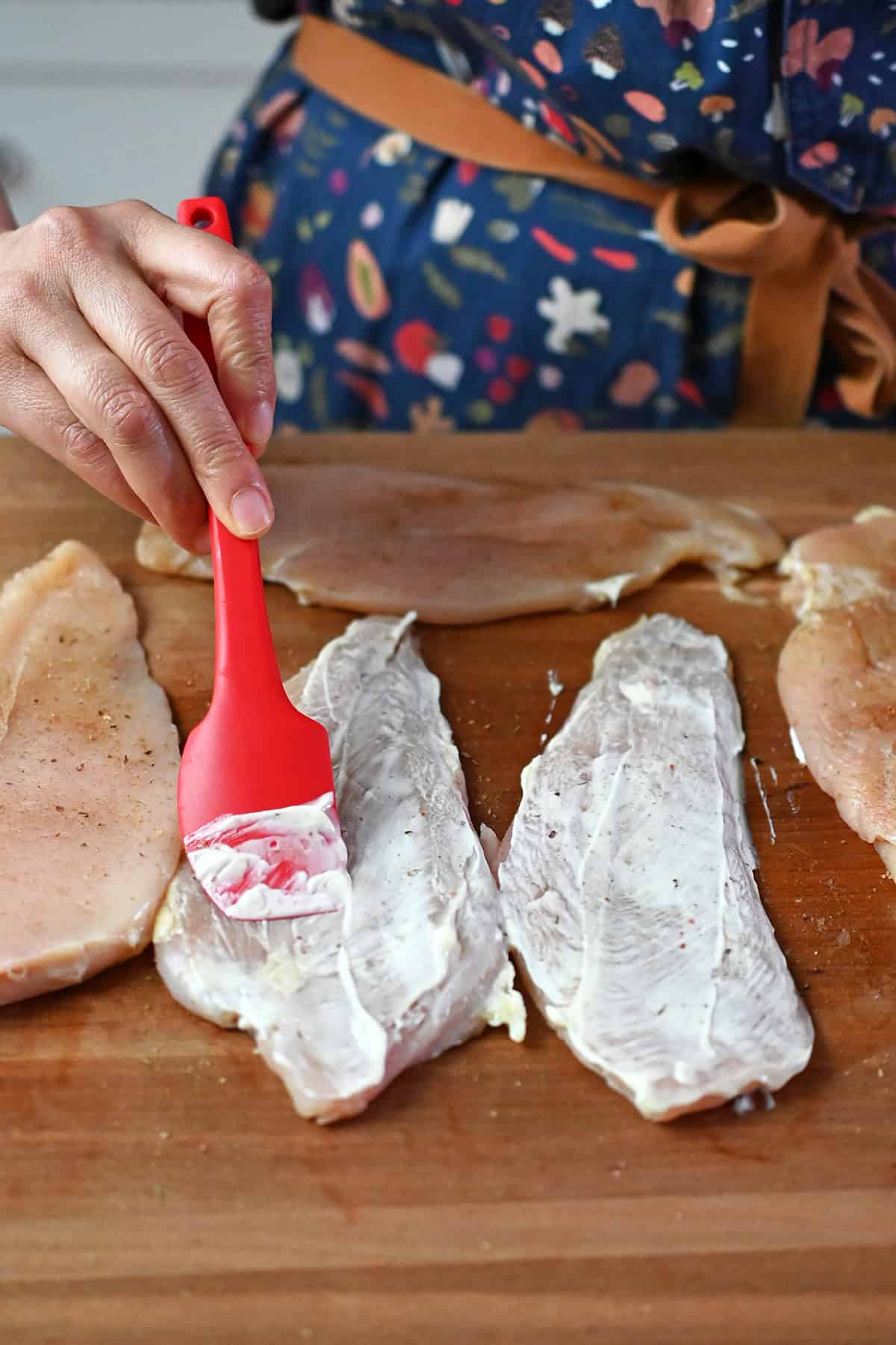 Brushing on a thin layer of mayonnaise on thinly cut raw boneless skinless chicken breasts with a small red spatula.