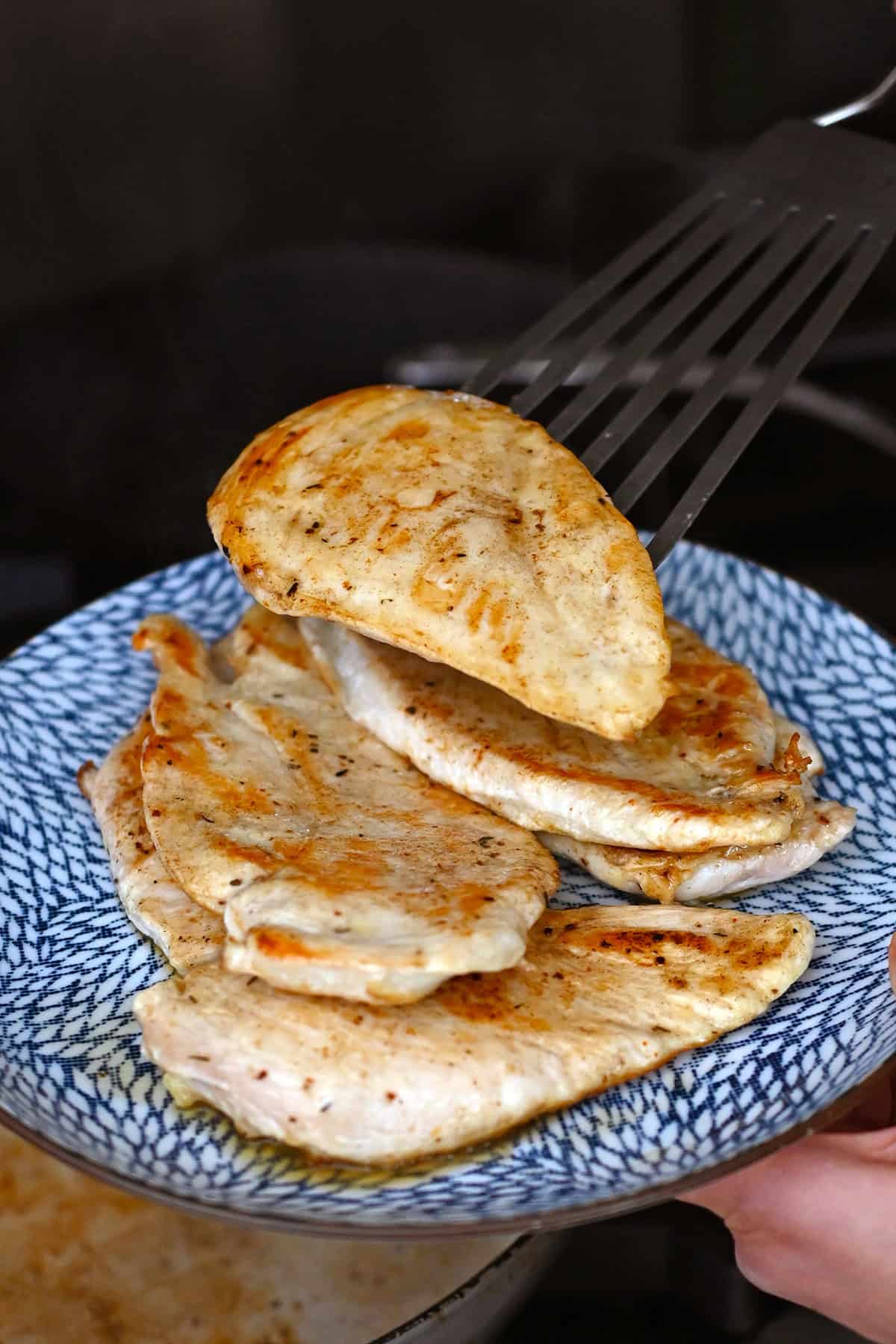 A person is using a metal fish spatula to transfer a golden brown chicken breast from a skillet onto a blue plate with other cooked chicken breasts.