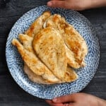Two hands are holding a blue plate filled with golden brown, perfectly cooked boneless, skinless chicken breasts.