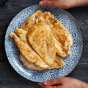 Two hands are holding a blue plate filled with golden brown, perfectly cooked boneless, skinless chicken breasts.